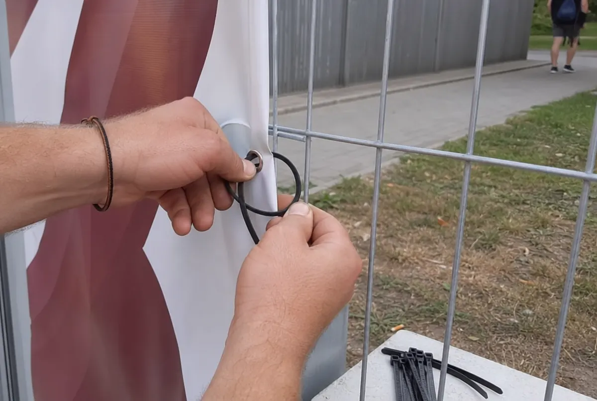 Mains fixant une bâche par l’œillet à un panneau de clôture avec un tendeur élastique