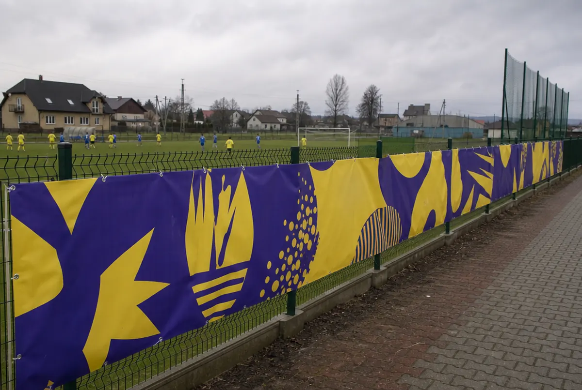Frontlit-Banner mit gelb-violetter Grafik am Maschendrahtzaun eines Sportplatzes
