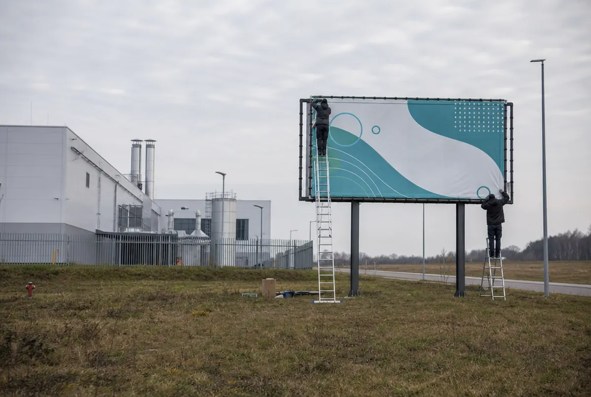 Installers tensioning a banner onto a company's own billboard frame by the road