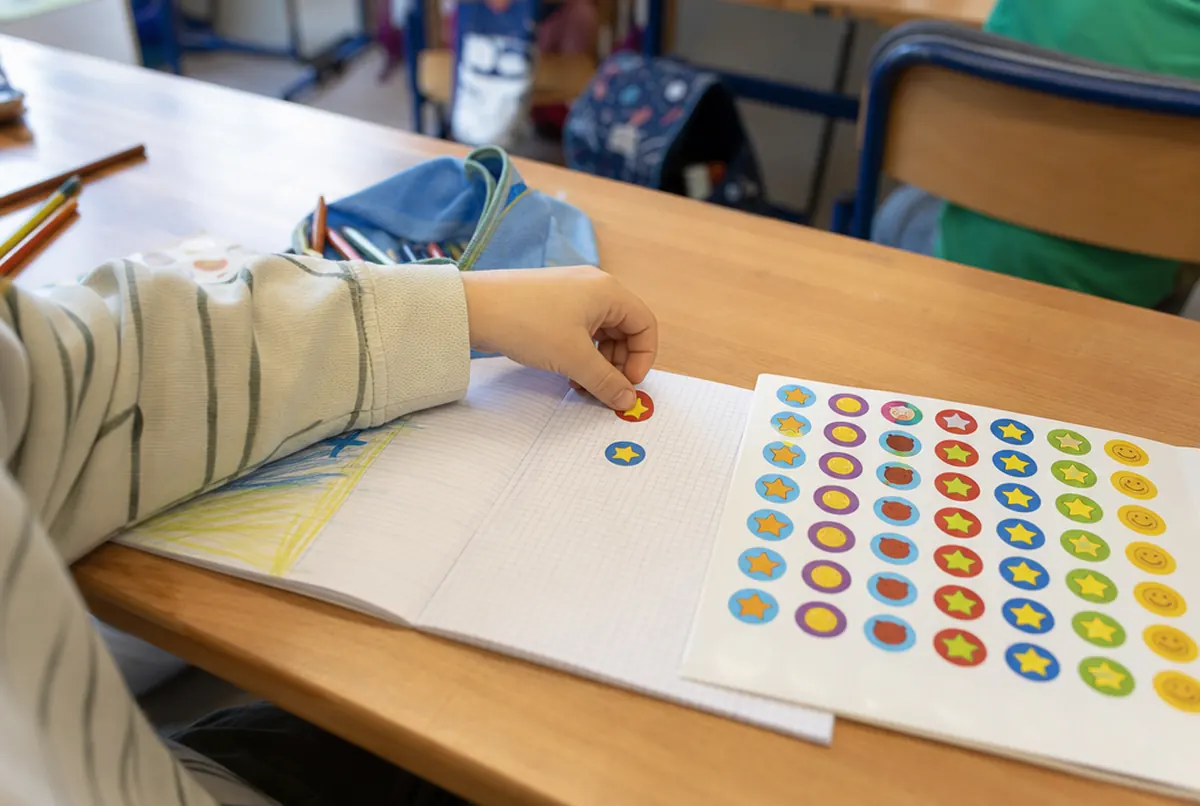 Child's hand placing a reward sticker from a colourful sheet in a school notebook