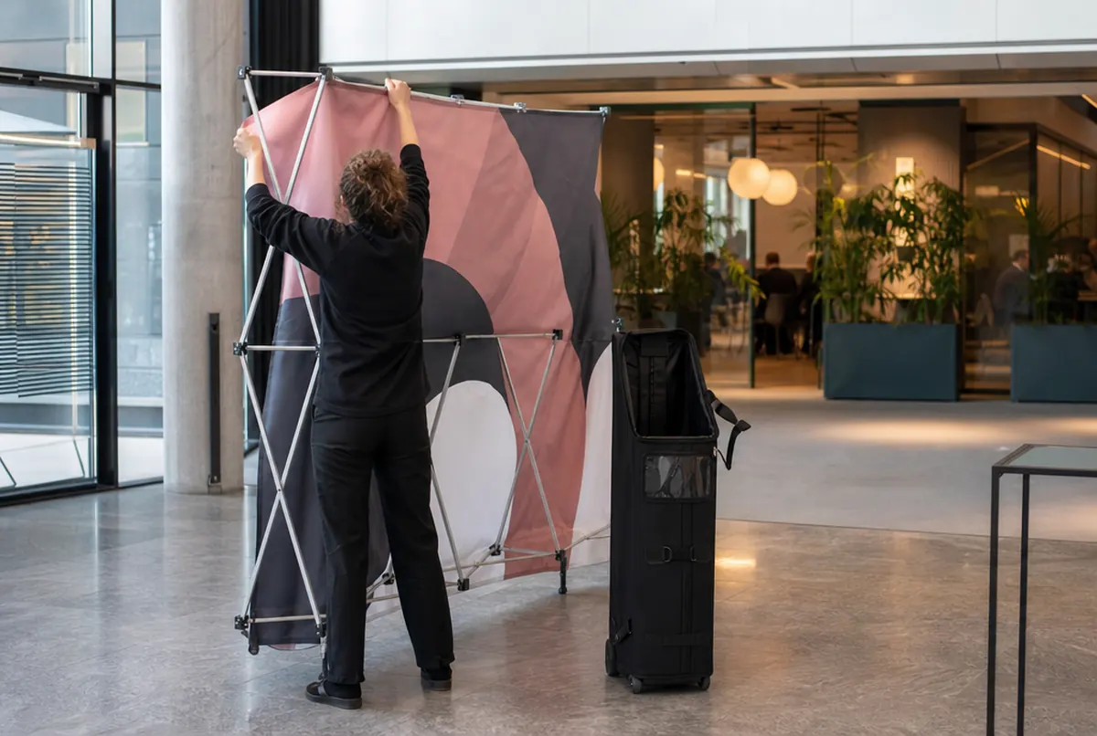 Unfolding a hop-up wall with its fabric attached next to a wheeled transport bag