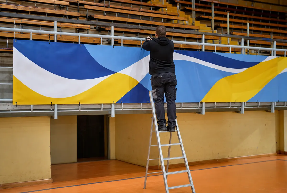 Ein Monteur auf der Leiter befestigt ein blau-gelbes Wellenbanner mit Kabelbindern am Tribünengeländer der Halle