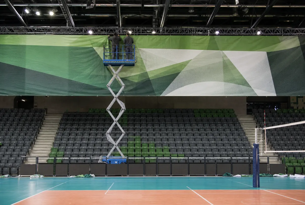 Montage von der Scherenbühne: zwei Monteure hängen ein großes grün-weißes Mesh-Banner unter das Hallendach