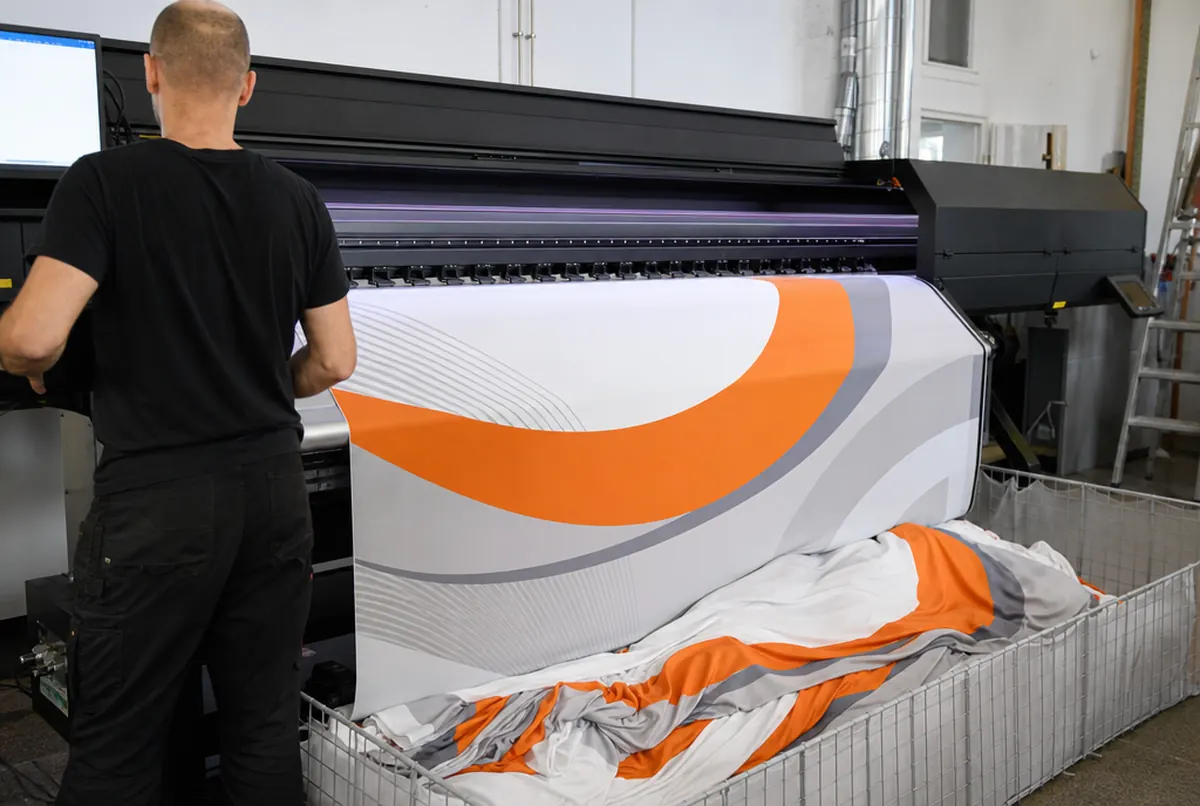 Ein Sublimationsdrucker druckt eine Textilgrafik mit orange-grauen Bögen, der Stoff fällt in einen Korb