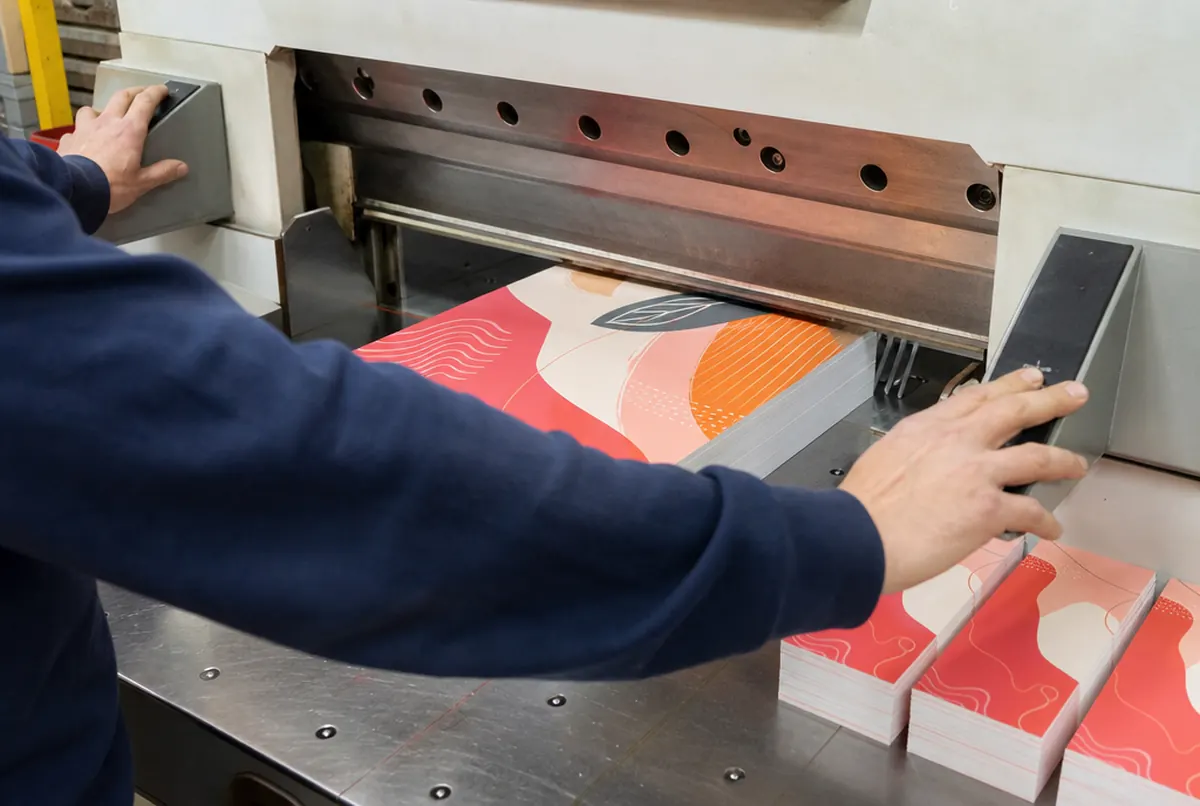 A paper guillotine trims a tall stack of flyers in coral and orange shapes, with cut bundles lined up beside it