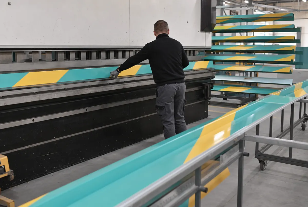 Produktionshalle: Dachfries-Paneele aus Verbundplatte mit türkis-gelben Diagonalstreifen werden abgekantet