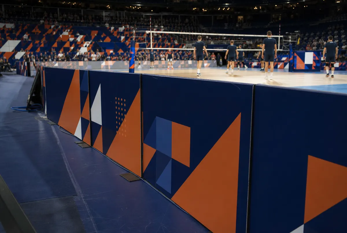 Spielfeldbanden mit dunkelblau-orangem geometrischem Muster an einem Volleyballfeld, dahinter Aufwärmen der Spieler