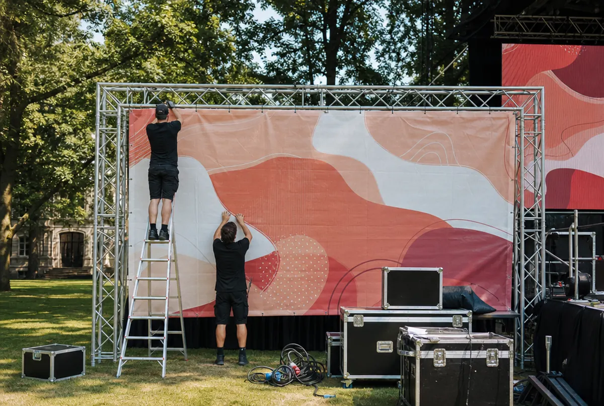 Team befestigt ein korallenrot bedrucktes Mesh-Banner mit Kabelbindern an der Traverse einer Freilichtbühne