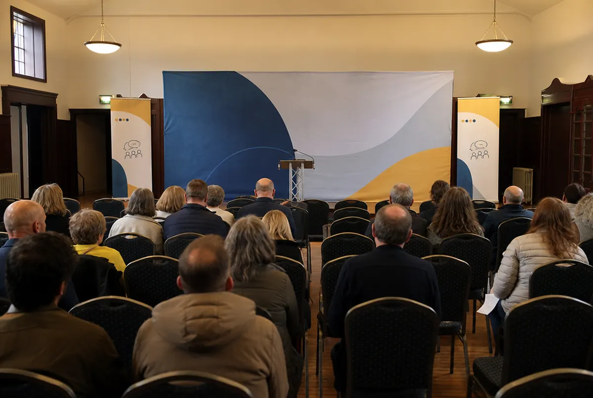 Sala spotkania z mieszkańcami z dwoma roll-upami i ścianką z abstrakcyjną grafiką, publiczność widziana od tyłu